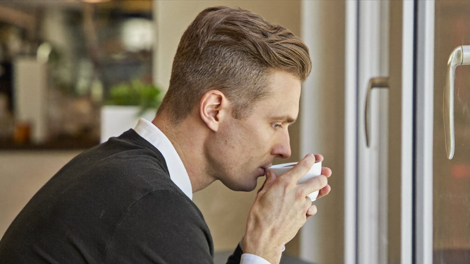 Man with neatly styled hair sipping coffee at a cafe window, deep in thought