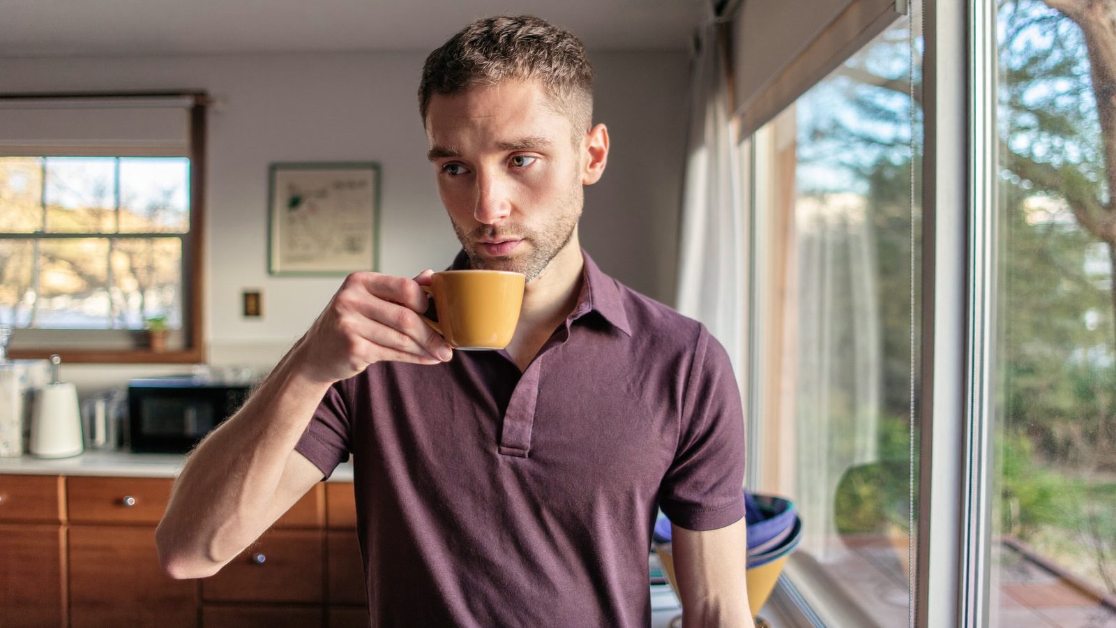 Man with short hair drinking coffee by a kitchen window in the morning