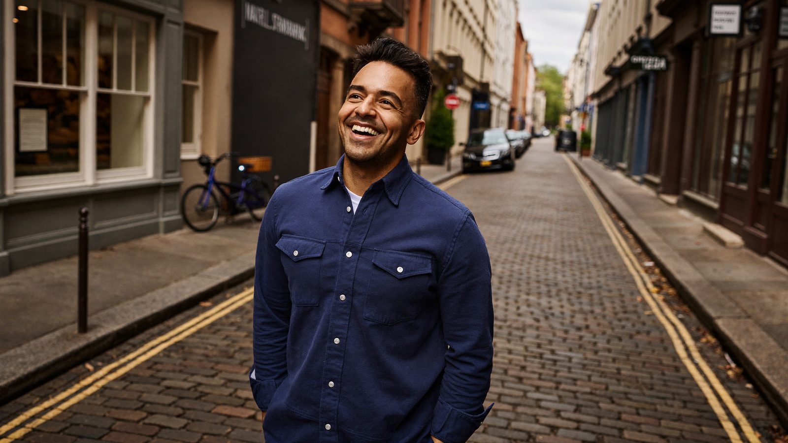 Smiling man in a navy shirt standing on a narrow cobblestone street lined with brick buildings