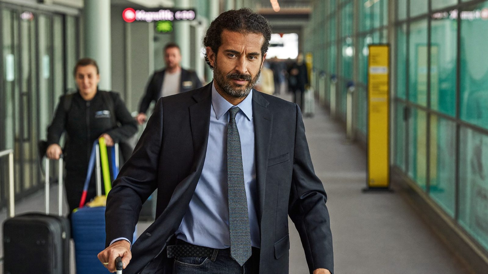 Man in a suit walking through an airport terminal with travelers and luggage behind him