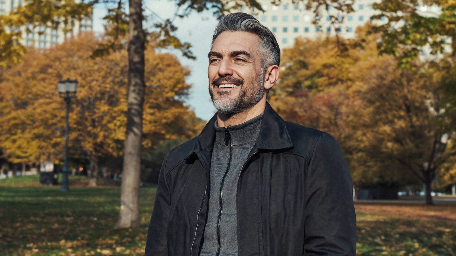 Smiling man in his 50s with full grey-flecked hair standing in a park in autumn