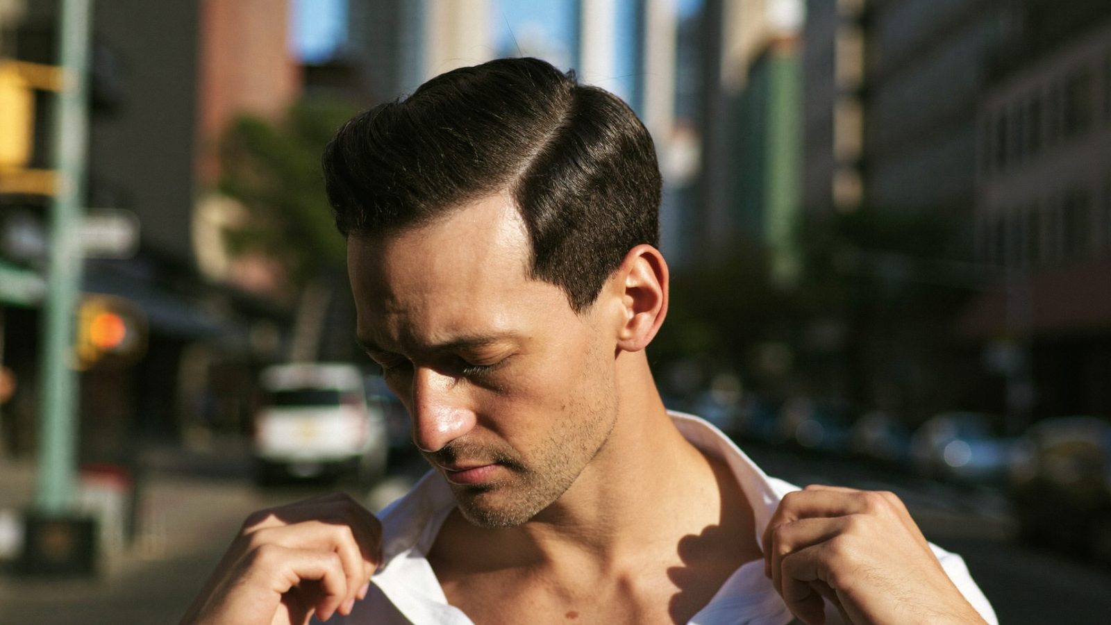 Man with neatly combed dark hair adjusting his shirt collar on a sunny city street